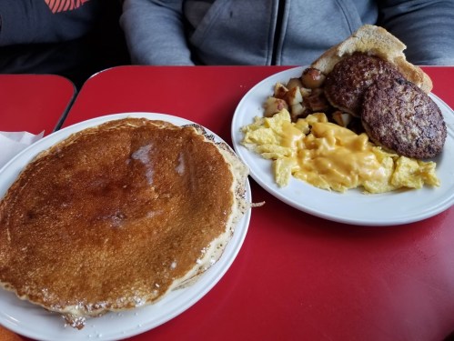 A plate of pancakes next to a plate with scrambled eggs, cheese, sausage patties, and diced potatoes.