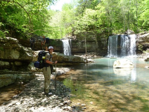 A hiker stands by a clear stream with two waterfalls in a lush green forest, surrounded by rocky terrain.