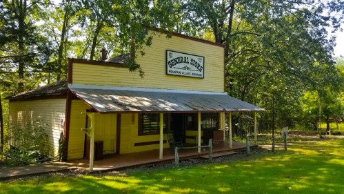 A yellow general store with a porch, surrounded by trees and greenery in a rural setting.