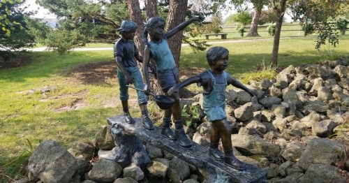 A bronze sculpture of three children playing by a rocky area, surrounded by greenery and trees.