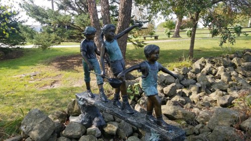 A bronze sculpture of three children playing on a rock, surrounded by trees and greenery.