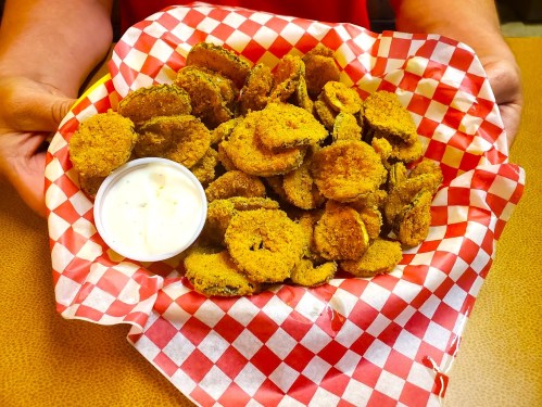 A person holds a basket of golden-brown fried pickles with a side of dipping sauce, lined with red and white checkered paper.