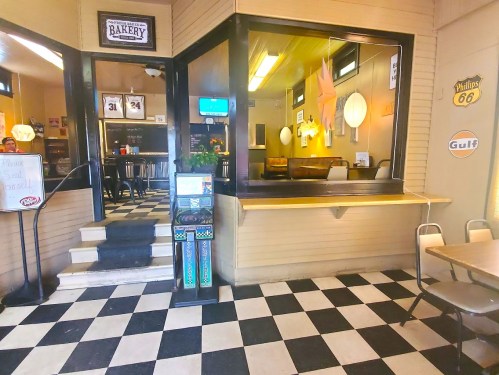 A cozy bakery with a checkered floor, featuring a counter, seating area, and a large window displaying the interior.