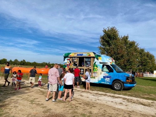 A colorful food truck with people and children gathered around it in a sunny outdoor setting.