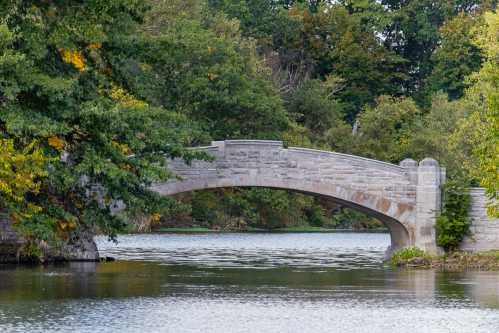 A stone arch bridge spans a calm river, surrounded by lush green trees and autumn foliage.