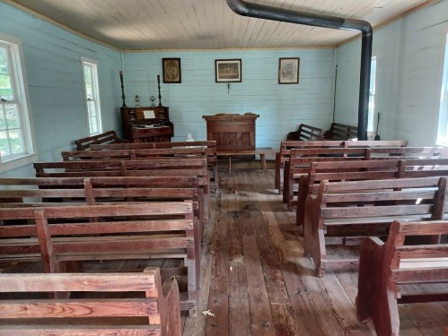 Interior of a rustic, empty church with wooden pews and a simple altar, featuring light blue walls and wooden flooring.