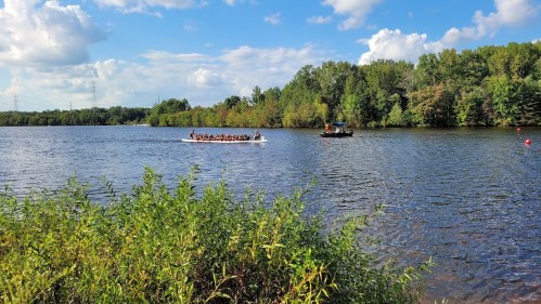 A group of people in a boat paddles on a calm lake surrounded by lush greenery and blue skies.