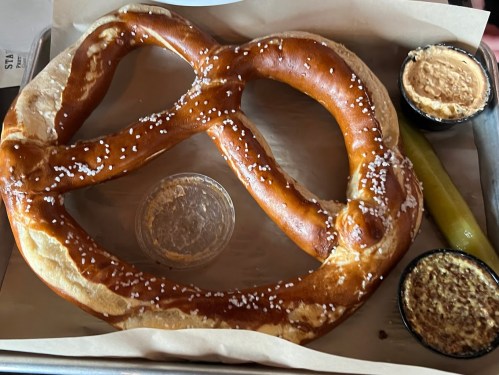 A large, soft pretzel with salt, served with mustard and cheese dip on a tray, alongside a pickle.