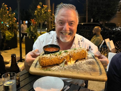 A smiling man holds a large, cheesy dish on a wooden platter, with a side of marinara sauce, outdoors at night.