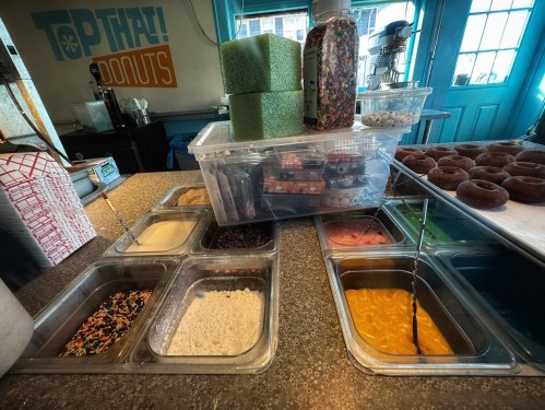 A countertop with various toppings for donuts, including sprinkles, icing, and sugar, alongside freshly made donuts.