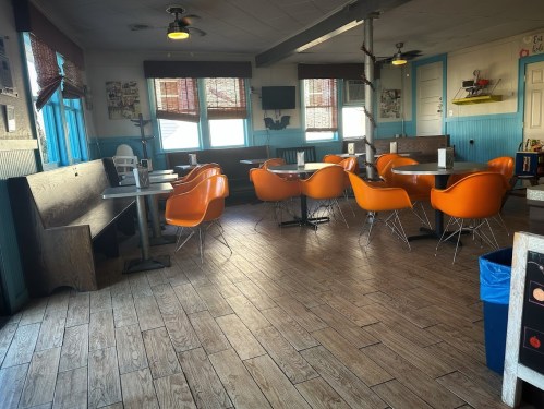 A cozy diner interior with orange chairs, wooden tables, and a blue color scheme, featuring large windows and a TV.