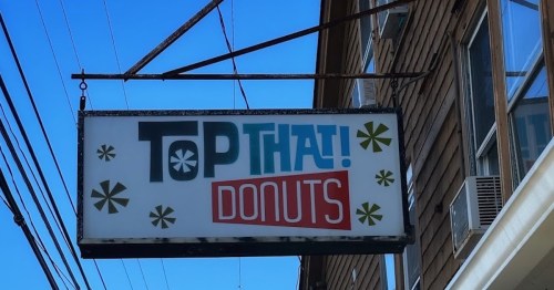 Sign for "Top That! Donuts" with colorful lettering and decorative snowflake designs against a clear blue sky.