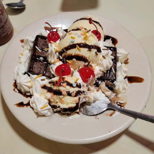 A dessert bowl with ice cream, chocolate sauce, whipped cream, brownies, and cherries, topped with colorful sprinkles.