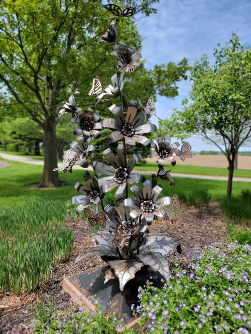 A metal sculpture of flowers and butterflies stands in a garden, surrounded by greenery and a clear blue sky.