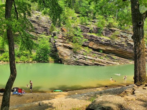 A serene river scene with people swimming and relaxing on the shore, surrounded by lush greenery and rocky cliffs.
