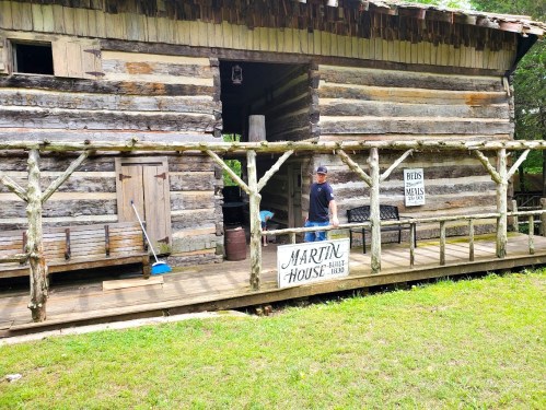 A rustic log cabin with a porch, featuring a sign that reads "Martin House 1810" and a person walking nearby.