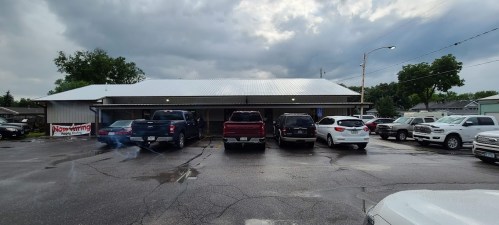 A parking lot with several vehicles in front of a building under a cloudy sky. Wet pavement reflects the surroundings.