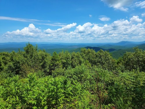 A panoramic view of lush green mountains under a bright blue sky with fluffy white clouds.