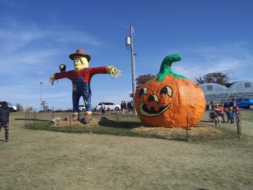 A large scarecrow and a giant pumpkin with a carved face stand in a field under a clear blue sky.