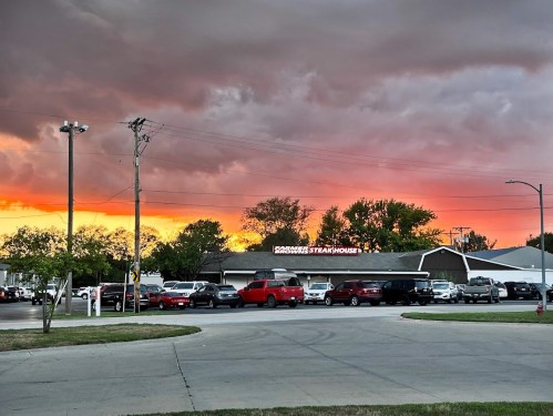 A vibrant sunset casts orange and pink hues over a busy parking lot in front of a steakhouse.