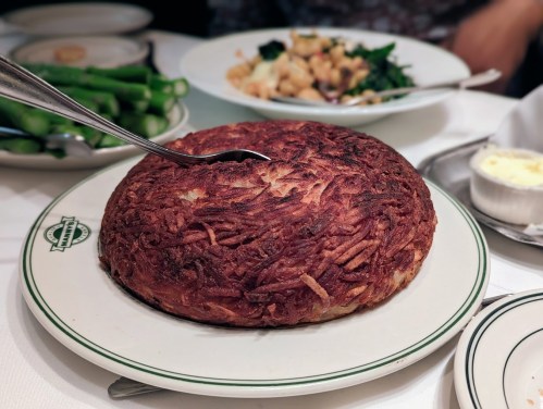 A golden-brown, crispy potato dish on a white plate, with a spoon resting on top, surrounded by other dishes.