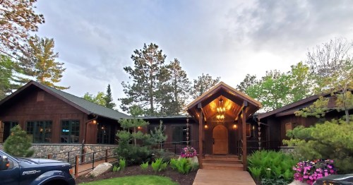 A rustic wooden cabin with a welcoming entrance, surrounded by trees and colorful flowers under a cloudy sky.