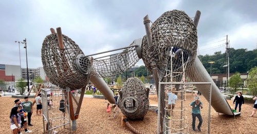 A modern playground with unique climbing structures and children playing, surrounded by families and greenery.