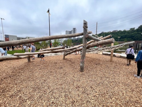 Children and adults play on a wooden climbing structure in a park, surrounded by gravel and greenery under a cloudy sky.