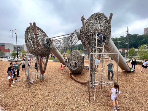A unique playground structure made of woven materials, with children playing and climbing on it, surrounded by gravel.