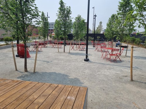 A spacious outdoor area with red chairs and tables surrounded by young trees and gravel pathways.