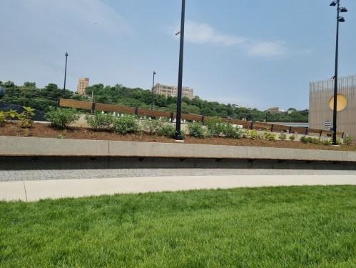 A landscaped area with greenery and a curved pathway, set against a backdrop of buildings and a clear blue sky.