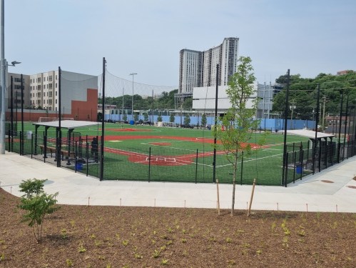 A baseball field surrounded by a fence, with buildings in the background and greenery in the foreground.