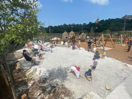 A playground scene with children playing in a sandy area, climbing structures, and adults supervising in a sunny environment.