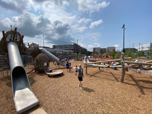 A vibrant playground with slides, climbing structures, and children playing under a sunny sky with scattered clouds.