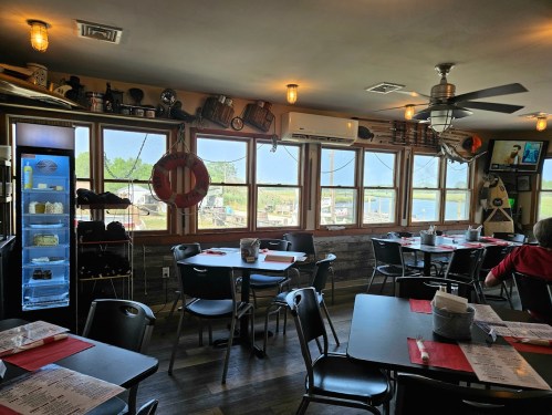 Cozy restaurant interior with tables, menus, and large windows overlooking a marina. Nautical decor enhances the ambiance.