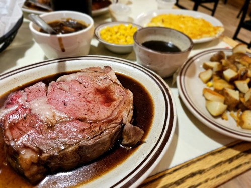 A plate featuring a large slice of prime rib in gravy, with sides of macaroni and cheese, potatoes, and soup.