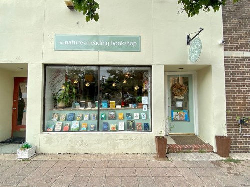 A cozy bookshop window displays a variety of books, with a welcoming entrance and greenery outside.