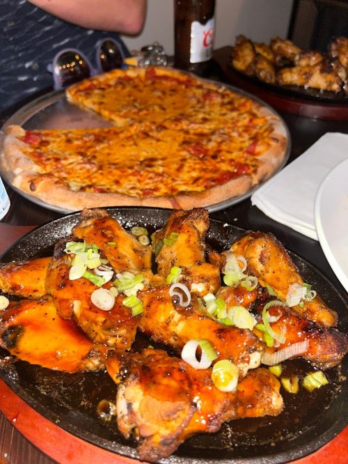 A plate of glazed chicken wings topped with green onions, with a pizza in the background.