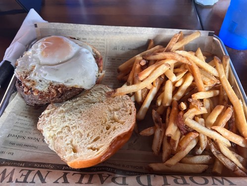 A burger with a fried egg on top, served with a side of crispy French fries on a tray.