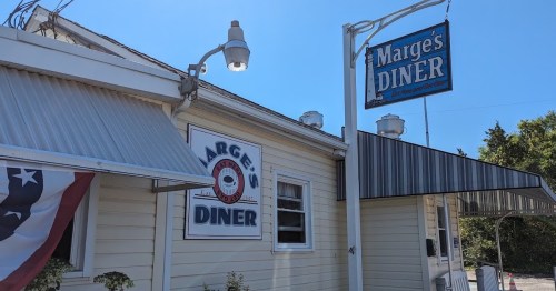 Exterior of Marge's Diner, featuring a sign and a flag, with a sunny sky in the background.
