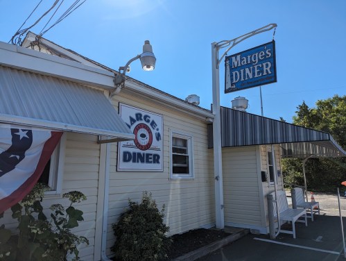 Exterior of Marge's Diner, featuring a sign and a white building with a porch and American flag decor.