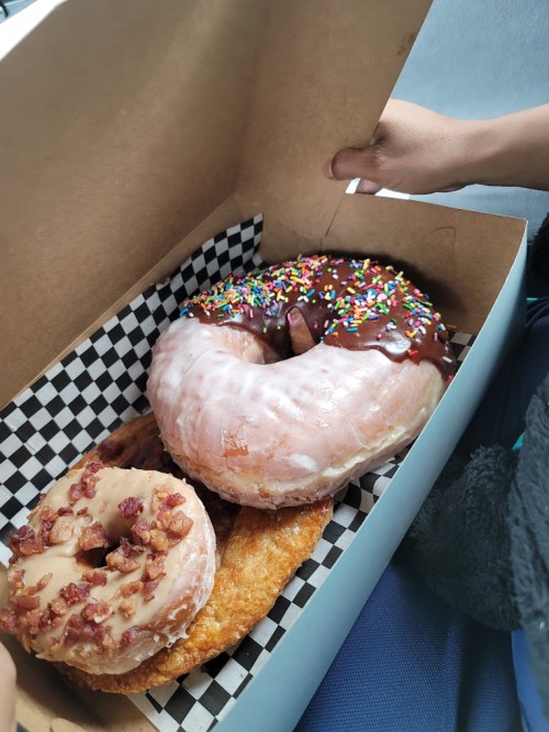 A box of assorted donuts: one with chocolate and sprinkles, one glazed, and one with bacon bits on top.