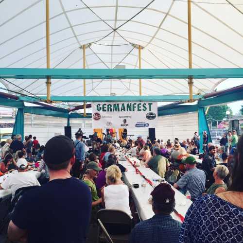 A large crowd gathers under a tent at Germanfest, enjoying food and festivities. Tables are set for dining.