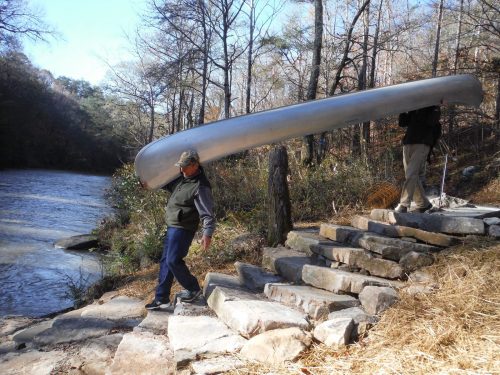 Two people carry a canoe along a rocky path by a river, surrounded by trees in a natural setting.