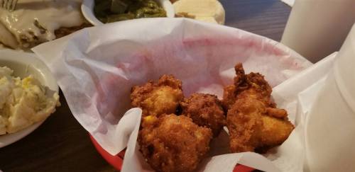 A basket of golden-brown fried food, possibly hushpuppies, on a table with sides of greens and other dishes in the background.