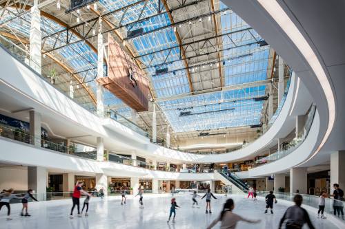 A spacious indoor ice skating rink with people skating, surrounded by modern architecture and large windows.