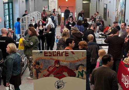 A busy event space filled with people browsing tables and displays, featuring comic-themed artwork and merchandise.