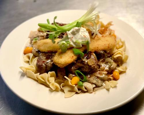 A plate of pasta topped with meat, vegetables, a dollop of sour cream, and crispy bread, garnished with green onions.
