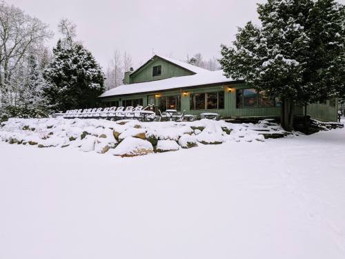 A green cabin surrounded by snow, with a stone wall and chairs lined up outside under a cloudy sky.
