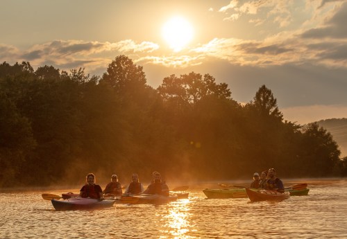 A group of kayakers paddles on a misty lake at sunset, surrounded by trees and a colorful sky.